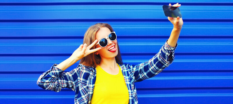 Portrait of cheerful young woman taking selfie by smartphone having fun wearing shirt on blue background