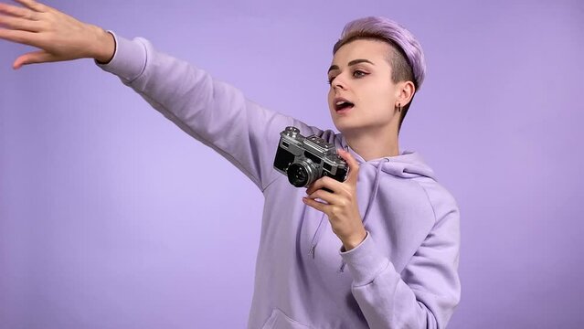 Positive young adult woman taking pictures with old-fashioned film camera, asking SB moving a little. Person generation Z using retro style device isolated on purple background. Synthwave concept