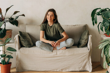 Portrait of a cheerful happy young female brunette sitting cross-legged on the sofa.