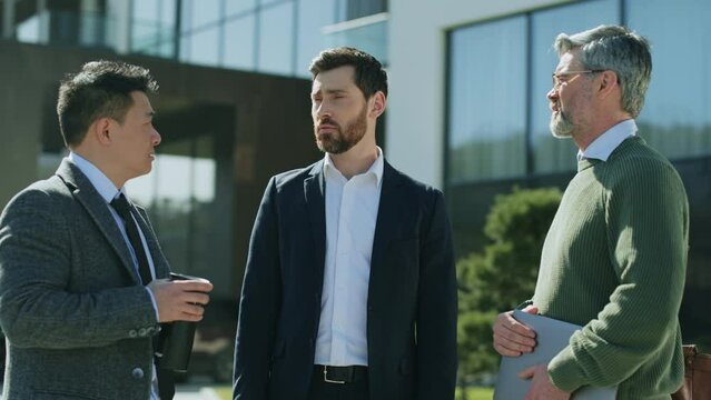 Group of serious confident multiethnic employees discussing working issues. Footage of businessmen dressed formal outfit gesturing and talking on background of business center. Marketing. Outdoor