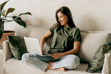 Young attractive brunette female student sitting on the sofa uses a laptop for online learning.