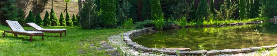 Fototapeta premium Lounge chairs on the backyard near the pond in a beautiful garden. Two empty sunbeds on the grass.