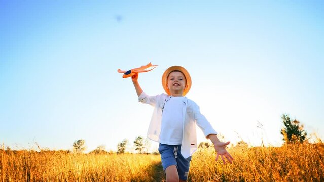 Happy baby boy runs and holds the airplane in his hand above his head against the wheat rural field and blue summer sky. Kid dream become pilot an astronaut spread his arms to sides and feeling free.