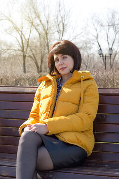 An adult brunette woman yellow jacket smiles gently sitting on a bench in the park.