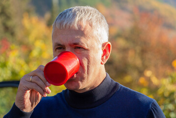 A man drinks tea or coffee from a red mug in nature.