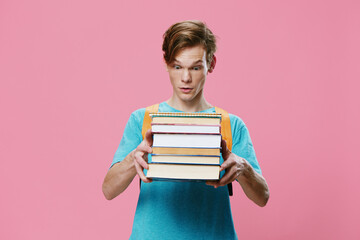 a red-haired student guy in a blue T-shirt and with an orange backpack on his back, holds a heavy stack of books in his hands, turning his face away from the camera. Horizontal photo with empty space