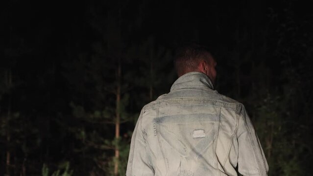 A preoccupied young man stands with his back turned at night in the forest. A man in the dark illuminated by a beam of light. Background out of focus with backlight. Nightlife, summer outdoors. 4K UHD