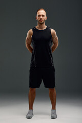 Full length portrait of young sporty guy in t-shirt and shorts standing on gray studio background. Serious millennial sportsman looking at camera. Healthy lifestyle and sports concept