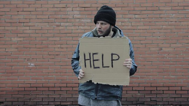 Homeless holds piece of cardboard with inscription Help. Refugee is at brick wall looking for job. Male tramp in dirty clothes, hat. Below poverty line. Bearded fugitive beggar. Immigrant. Drunkenness