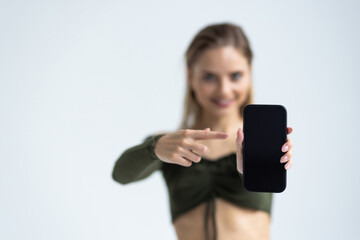 Naklejka premium Portrait of a joyful woman pointing finger at blank screen mobile phone and looking at camera isolated over white background