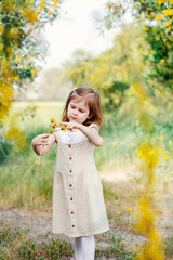 Cute toddler girl with long hairs wear casual dress walking, runing in field with green grass and blooming trees with yellow flowers.