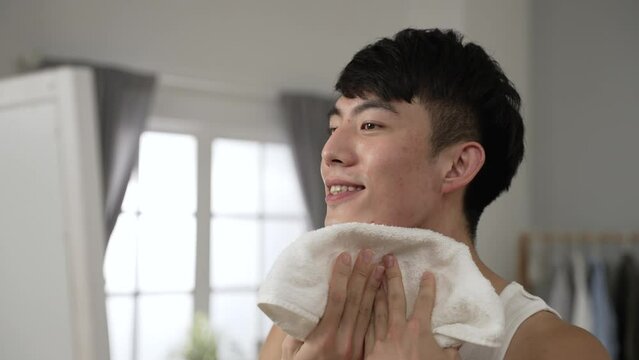 happy asian man in undershirt is looking at his reflection in the mirror and wiping face with a towel during daily skincare routine in the morning at home