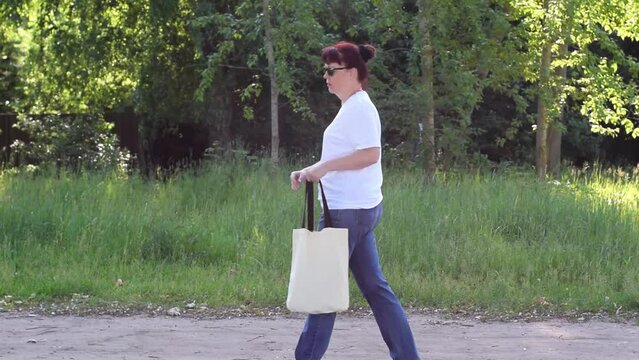 Mature adult woman with red hair wearing white T-shirt and jeans is walking carrying a white tote sack in summer. She stops to find something in the bag. Reusable bag made of sustainable materials