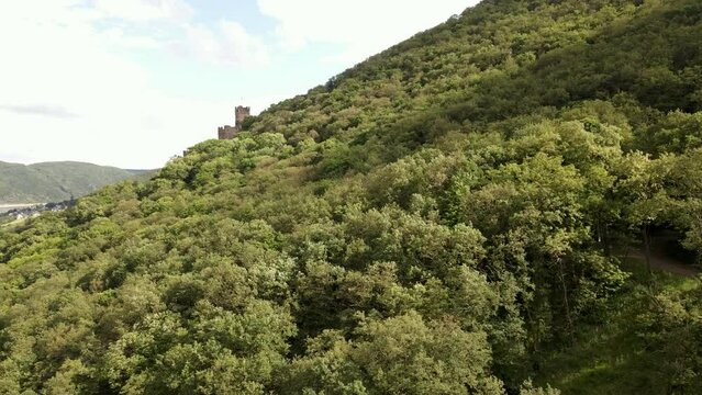 Ancient European hillside castle revealed behind a lush deciduous forest on a sunny day. Aerial ascending slider shot