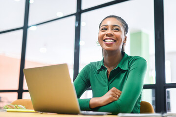 Inspired happy african-american female employee colleague enjoys her work, sitting on the workplace with laptop, looks away and loughts, successful black businesswoman in friendly office atmosphere