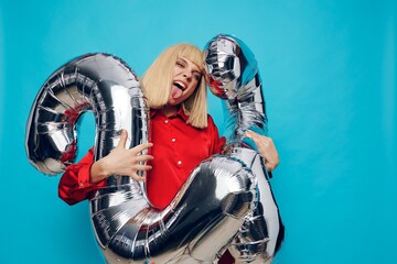 a bright, emotional, funny woman in a red shirt stands on a blue background and holds inflatable balloons in the shape of the number twenty-two in silver color showing her tongue to the camera