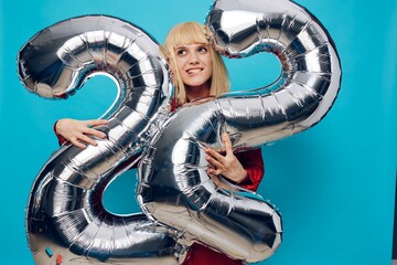 a woman in a red shirt stands showing her teeth on a blue background and holds inflatable balloons in the shape of the number twenty-two in silver color hugging them with her hands