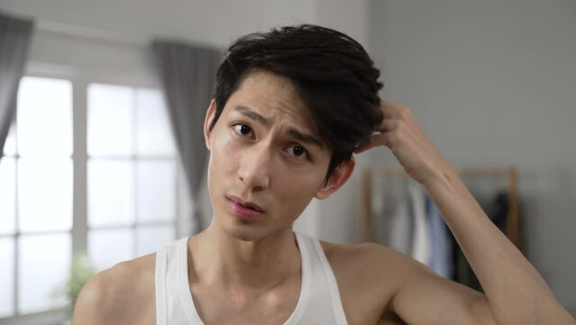 cheerful asian man looking at the camera as mirror is combing his hair with hand and smiling while going through morning beauty routine in the bedroom at home.