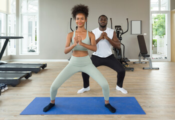 Obraz premium Young curly black hair instructor in sportswear leading exercise class how to do bodyweight squat. Cardio machines are on the background at the gym.