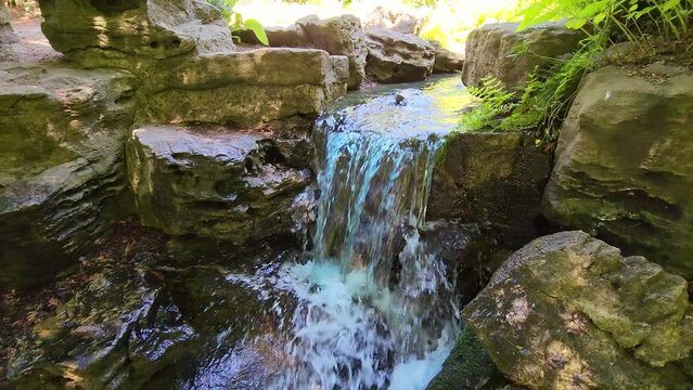 Video of a small waterfall, mossy rocks and flowing water stream. A small waterfall cascades over mossy rocks into a flowing stream, creating a tranquil natural scene.