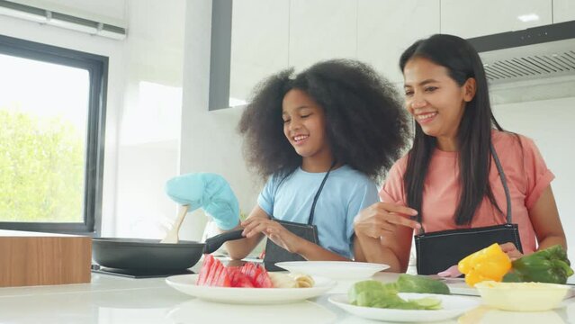 Cute young long curly hair African girl cooking food in kitchen, happy enjoy eat food, young kid do breakfast with her Asian mom at home