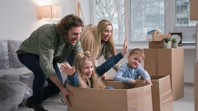 cheerful young parents running with boxes with daughter and son ride inside, family homeowners playing having fun on relocation