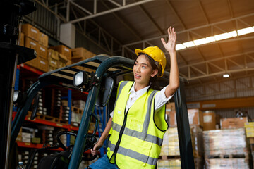 Asian women staff worker in uniform and yellow helmet in warehouse, smiling and waving somebody greetings.