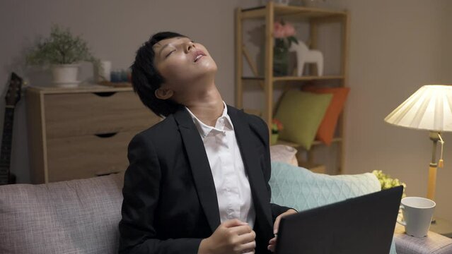 tired asian woman manager sitting on couch with a computer is exercising her stiff neck with a shoulder shrug while working overtime in the living room at home.
