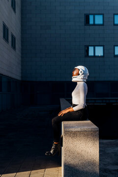 Businesswoman wearing space helmet sitting on wall by office building