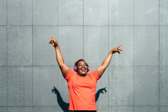 Cheerful woman with arms raised enjoying music in front of wall on sunny day