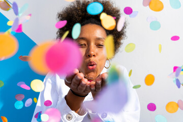 Young woman blowing multi colored confetti in front of wall on sunny day