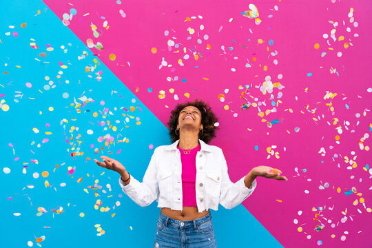 Happy woman with Afro hairstyle standing amidst confetti in front of pink and blue wall