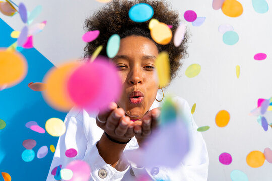 Young woman blowing multi colored confetti in front of wall on sunny day