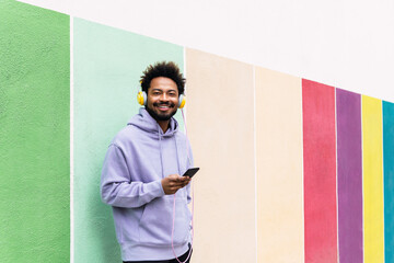 Smiling man holding smart phone listening music through headphones in front of colorful wall