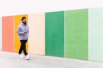Man holding mobile phone listening music through headphones walking on footpath by colorful wall