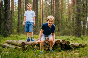 Two young small cute adorable playful caucasian school boys enjoy having fun walking at summer forest jumping and balancing on log tree outdoor. Healthy outside nature family activity.