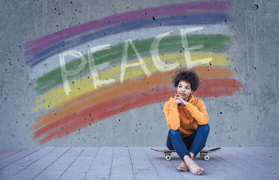Young woman sitting on skateboard in front of rainbow flag with text Peace