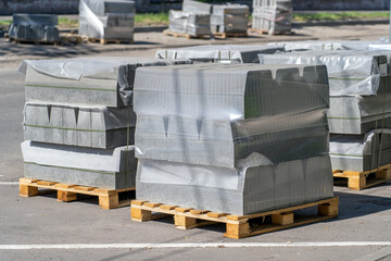 Stack of grey concrete kerbs on wooden pallet wrapped with film stand at site of repair of street. Road works, sidewalk repair