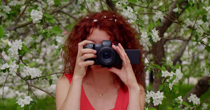Woman photographer in a red dress in a blooming park makes a photo with a camera. Concept tourist on vacation makes travel footage.