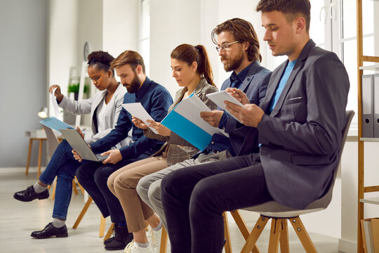 Job seekers waiting for job interview. Diverse group of people sitting in row in office, reading their CVs, using laptops and waiting for their turn to be interviewed. Business recruitment concept