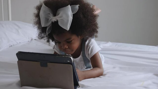 Pre-school-aged African female child lying on bed, watching and using digital tablet alone at home. Juvenile, digital device, learning and lifestyle concepts.