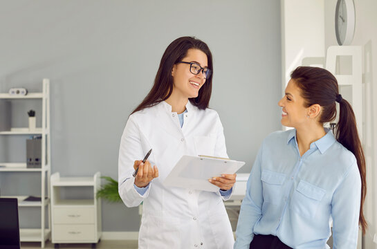 Friendly Cheerful Nurse Or Doctor At Modern Clinic Talking To Patient. Happy Physician Or Gynaecologist Holding Clipboard, Smiling And Giving Medical Consultation To Young Woman. Health Concept