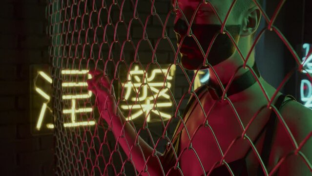 Tilting up of girl with shaved head, black paint on face, in white contact lenses holding netting fence in studio with neon lights in shapes of hieroglyphs meaning nirvana and western paradise