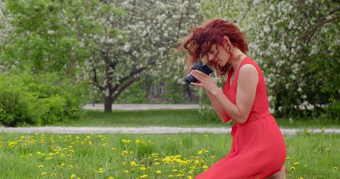 Woman photographer in a red dress in a blooming park makes a photo with a camera. Concept tourist on vacation makes travel footage.