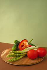Vegetables lie on a wooden board: tomatoes, asparagus, cucumbers, red bell peppers. brown, light green background. place for text.