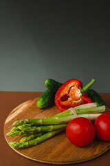 Vegetables lie on a wooden board: tomatoes, asparagus, cucumbers, red bell peppers. brown, dark gray background. place for text.