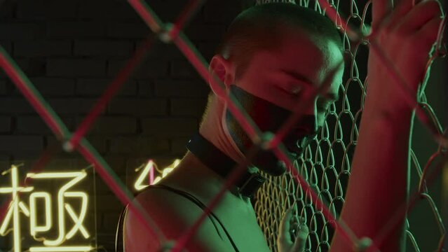 Shoulder-up of woman with black paint on face, in white contact lenses holding netting fence, looking on camera in studio with neon lights in shapes of hieroglyphs meaning nirvana and western paradise
