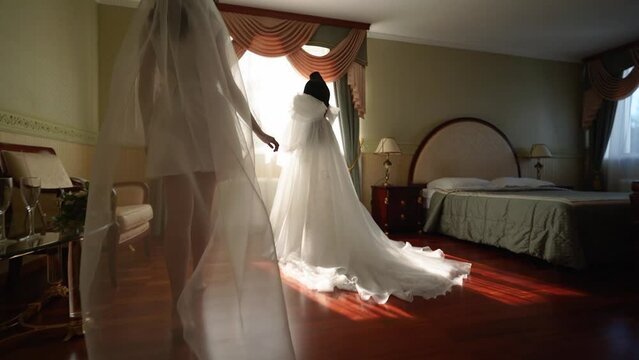 Bride enters the room in long bridal veil, strokes and touches her wedding dress with her hands. Woman admires her white dress.