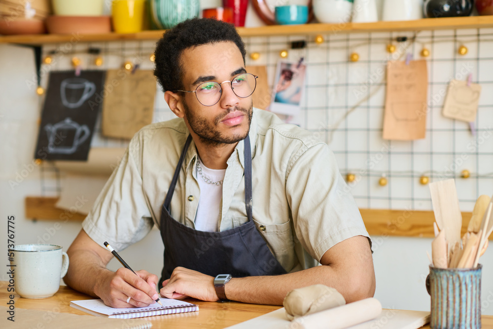Poster young pensive black man in workwear looking aside while sitting by workplace and drawing sketch of n