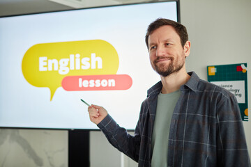 Waist up portrait of bearded male teacher pointing at digital whiteboard with English lesson text, copy space